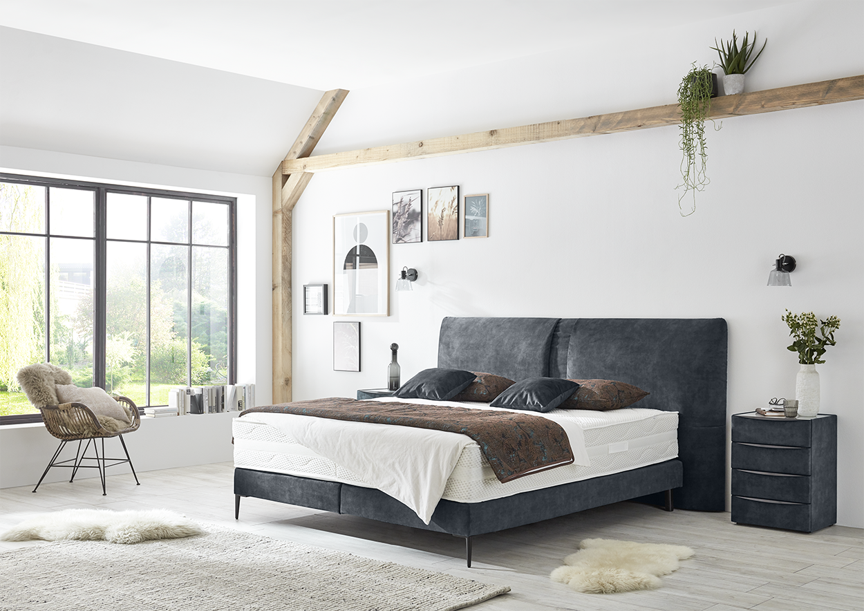 Modern eingerichtetes Schlafzimmer mit Polsterbett in Dunkelgrau, Nachttisch und grossem Fenster mit Blick ins Grüne. plaintext Kopieren Bearbeiten